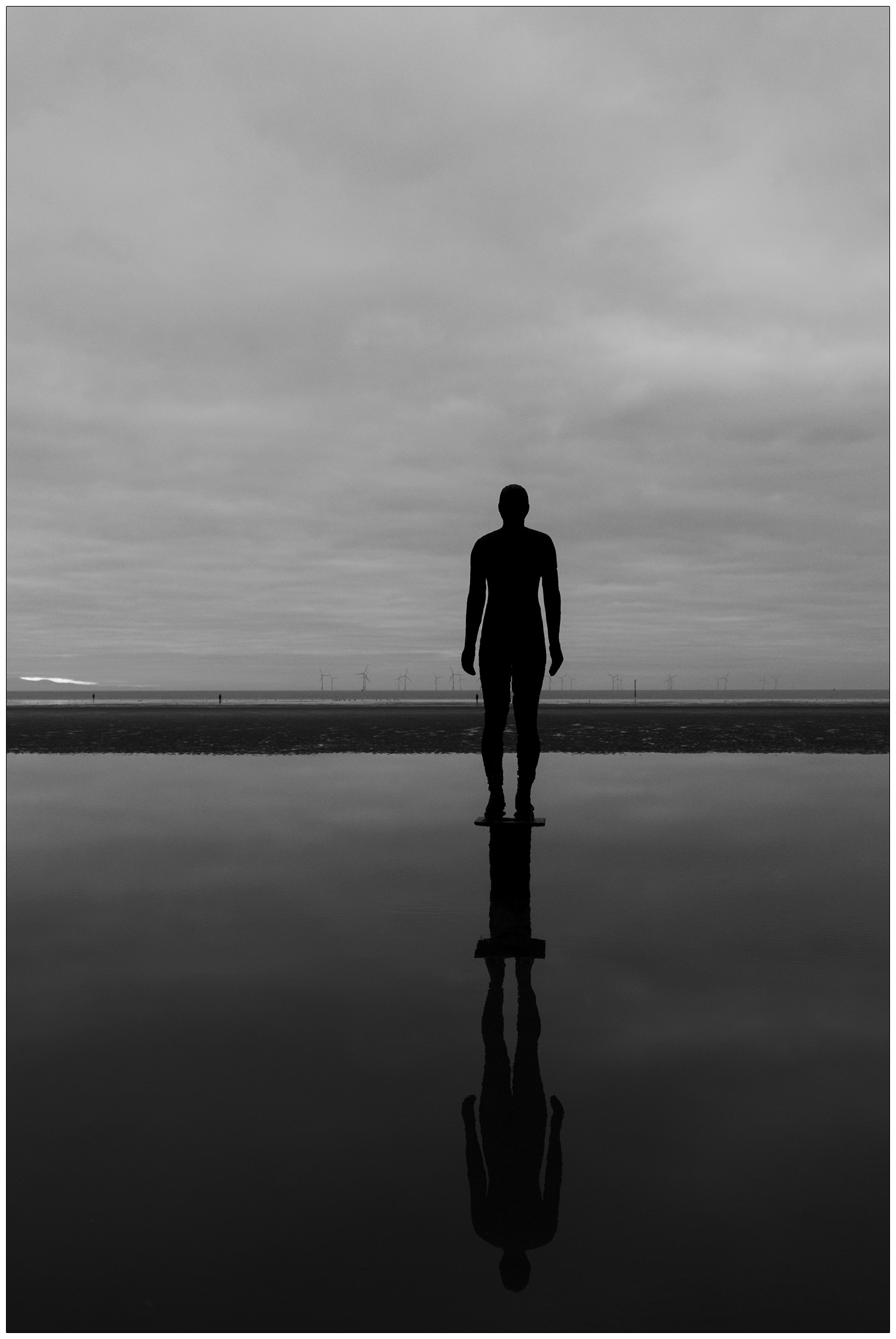 black and white photo of Another Place sculpture by Antony Gormley. The cast iron figure is reflected in the water and looks out at wind turbines on Crosby beach