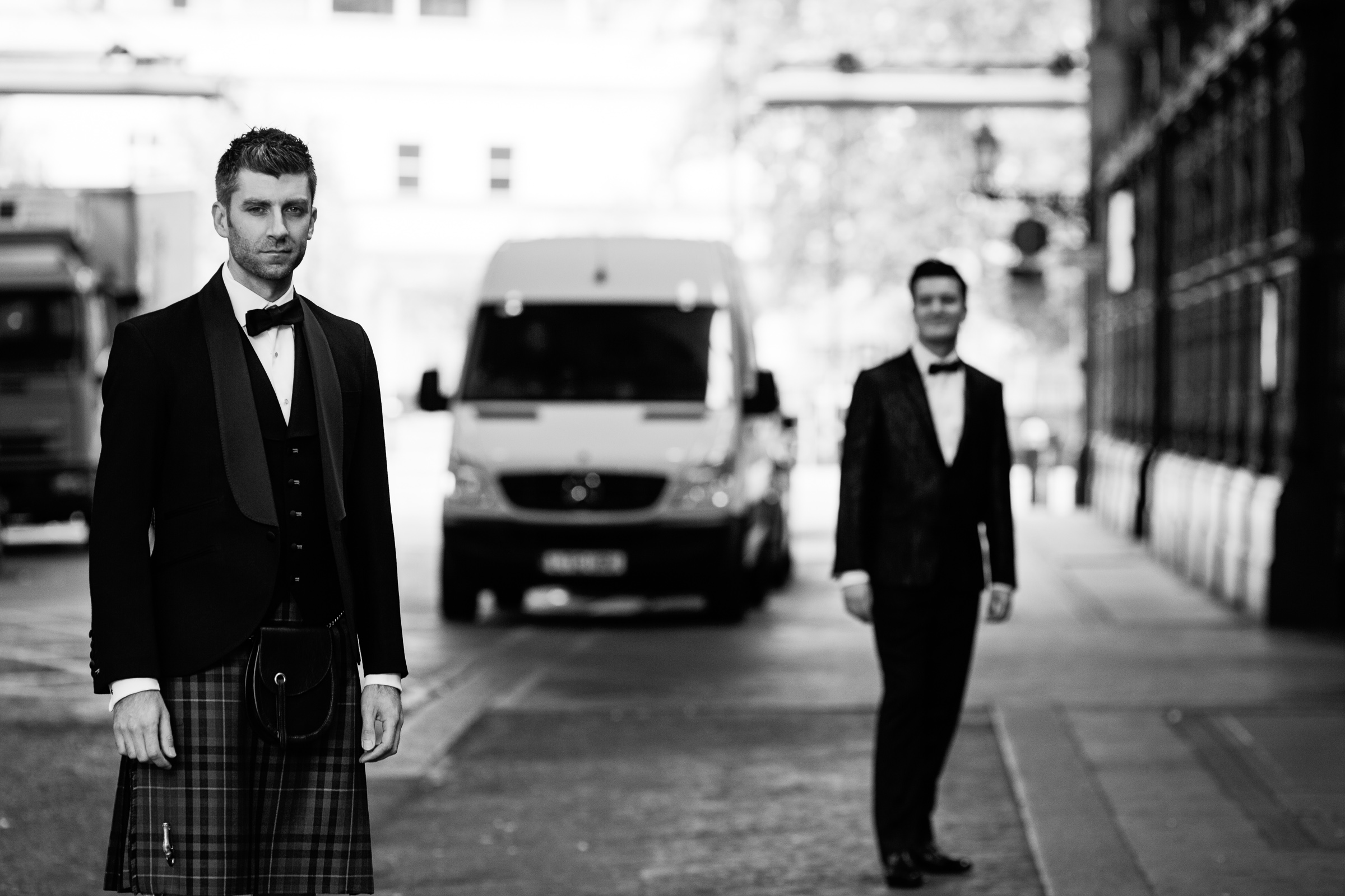 A portrait of two men on the way to their civil partnership ceremony at the Haberdashers' Hall in Smithfield. The man in the foreground is wearing kilt at bow tie. the ban behind is in black tie. There is a white van in the road.
