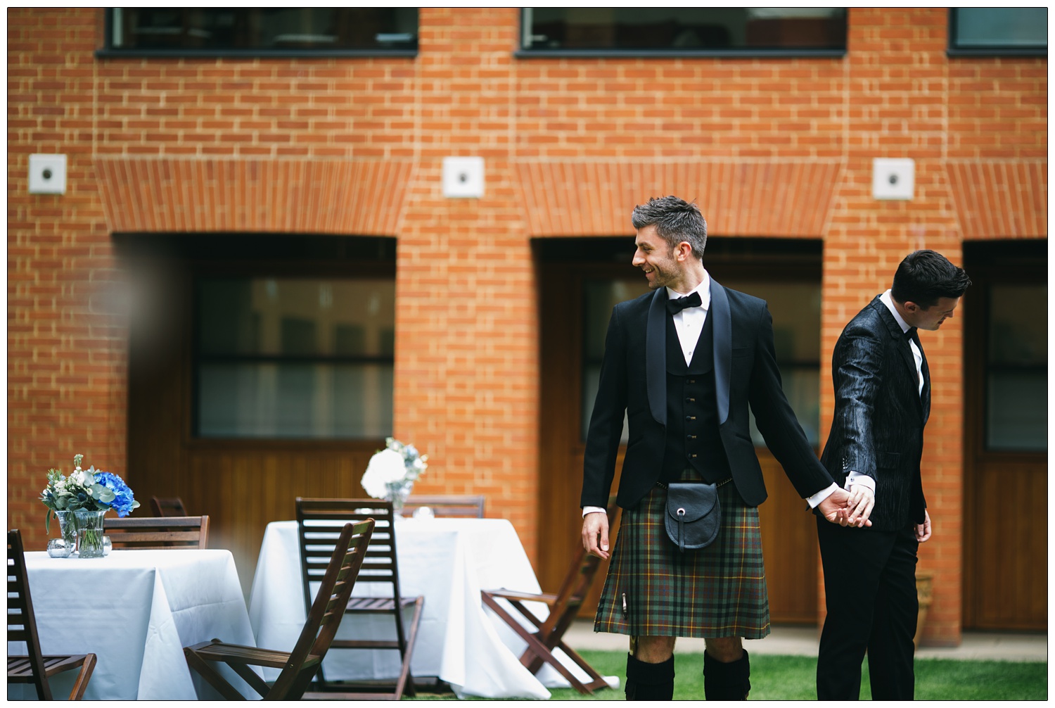 A man in a kilt and man in a suit are holding hands outside in a courtyard.