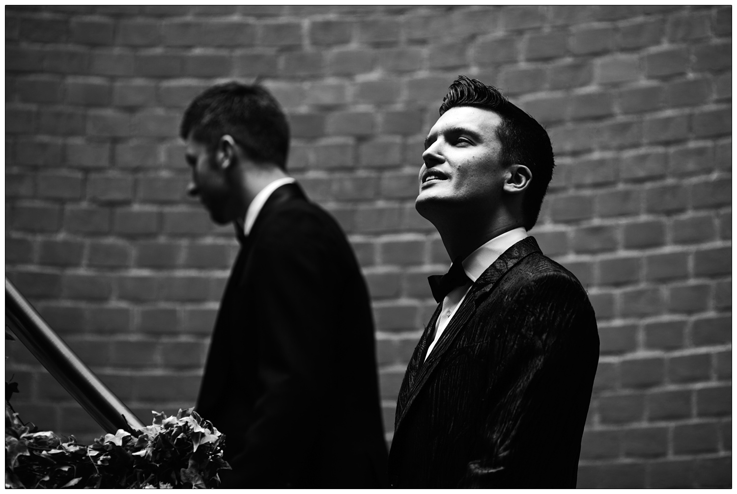 Two men walking up the stairs. The light from above is illuminating a man's face. He is wearing a black jacket and bow tie.