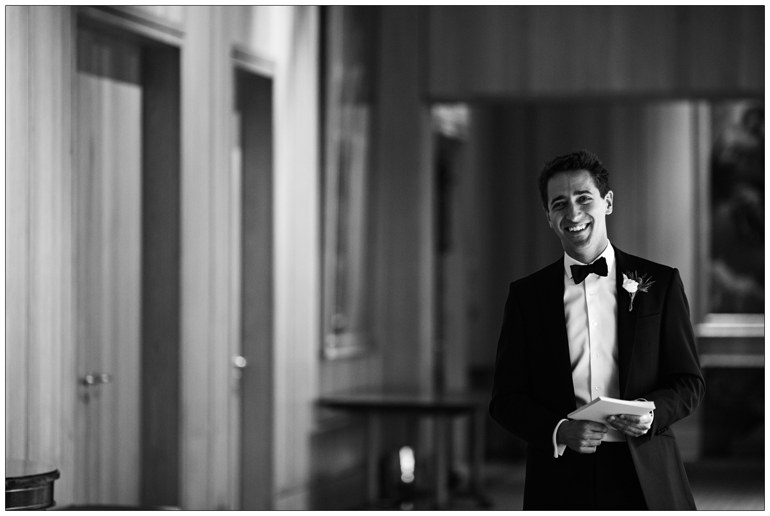 A man in a black suit and bow tie walks along holding some cards and smiling.