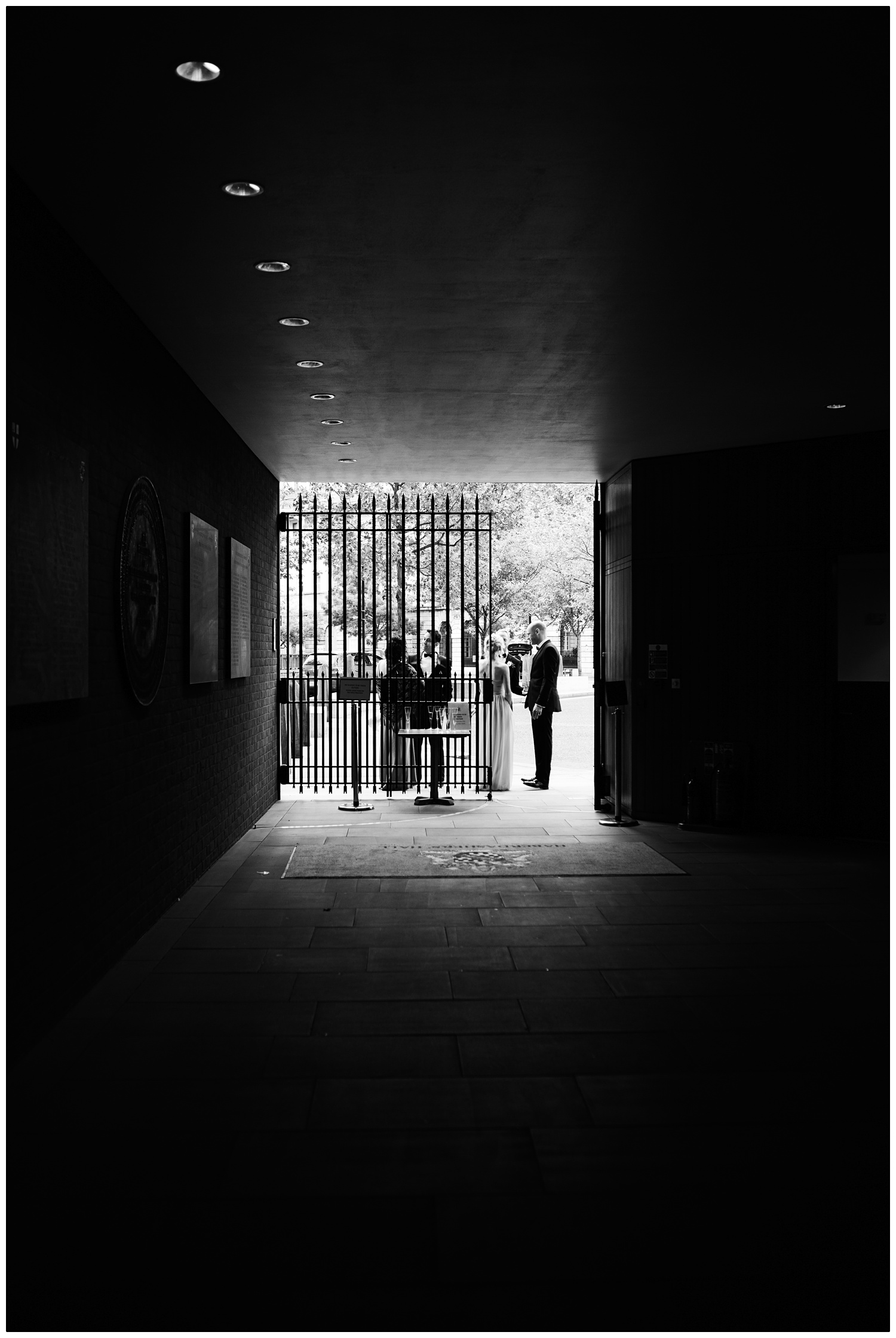 View down a corridor. There is a gate and wedding guests are outside.