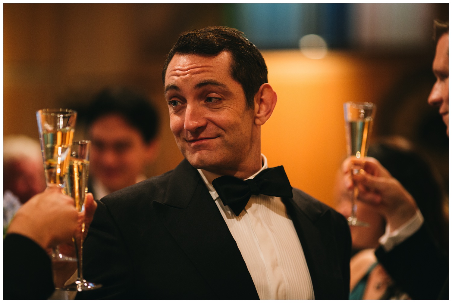A dapper looking man in a bow tie smiles while people raises their glasses.