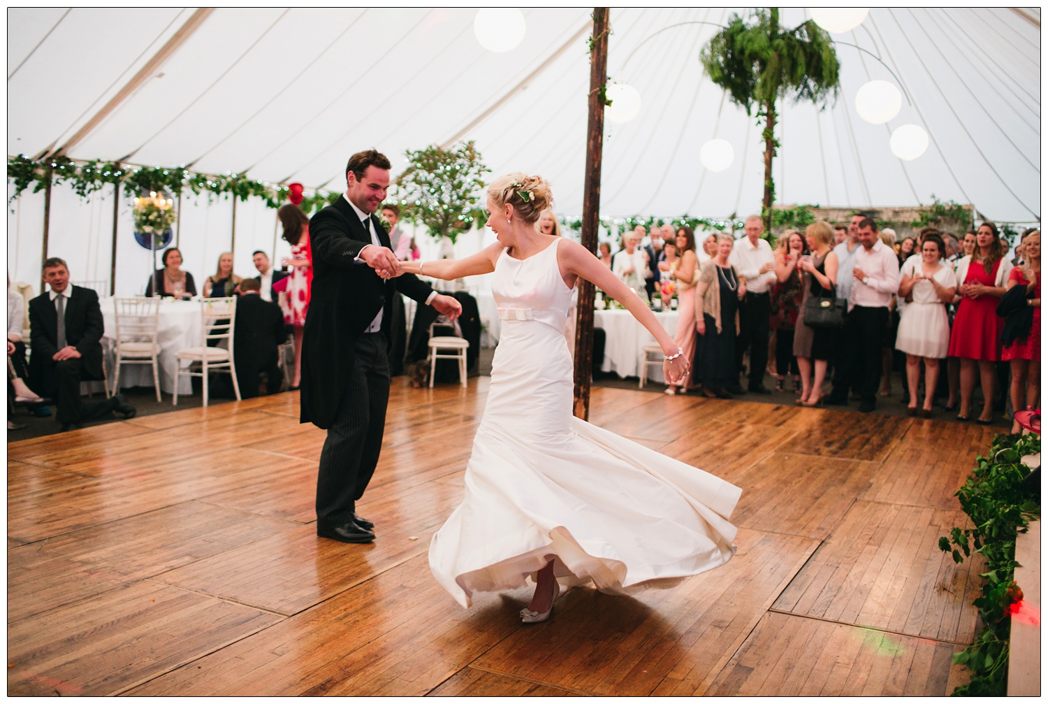 First dance in a marquee decorated with fairy lights and green foliage. The bride is twirling round.