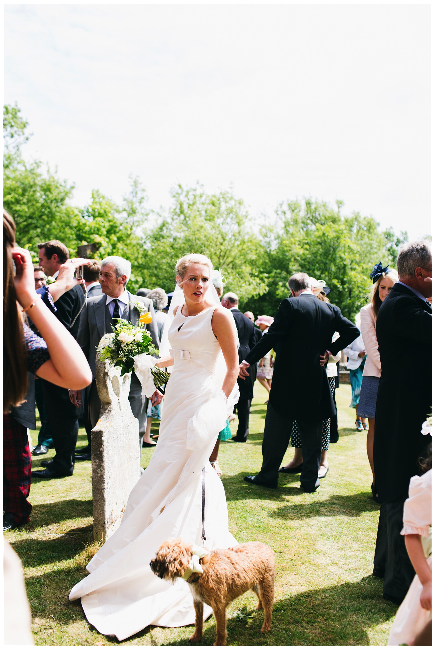 Bride is outside the St Thomas’ church in Bradwell-on-Sea. Her dog is next to her on a lead. A reportage style unposed wedding photograph in Essex.