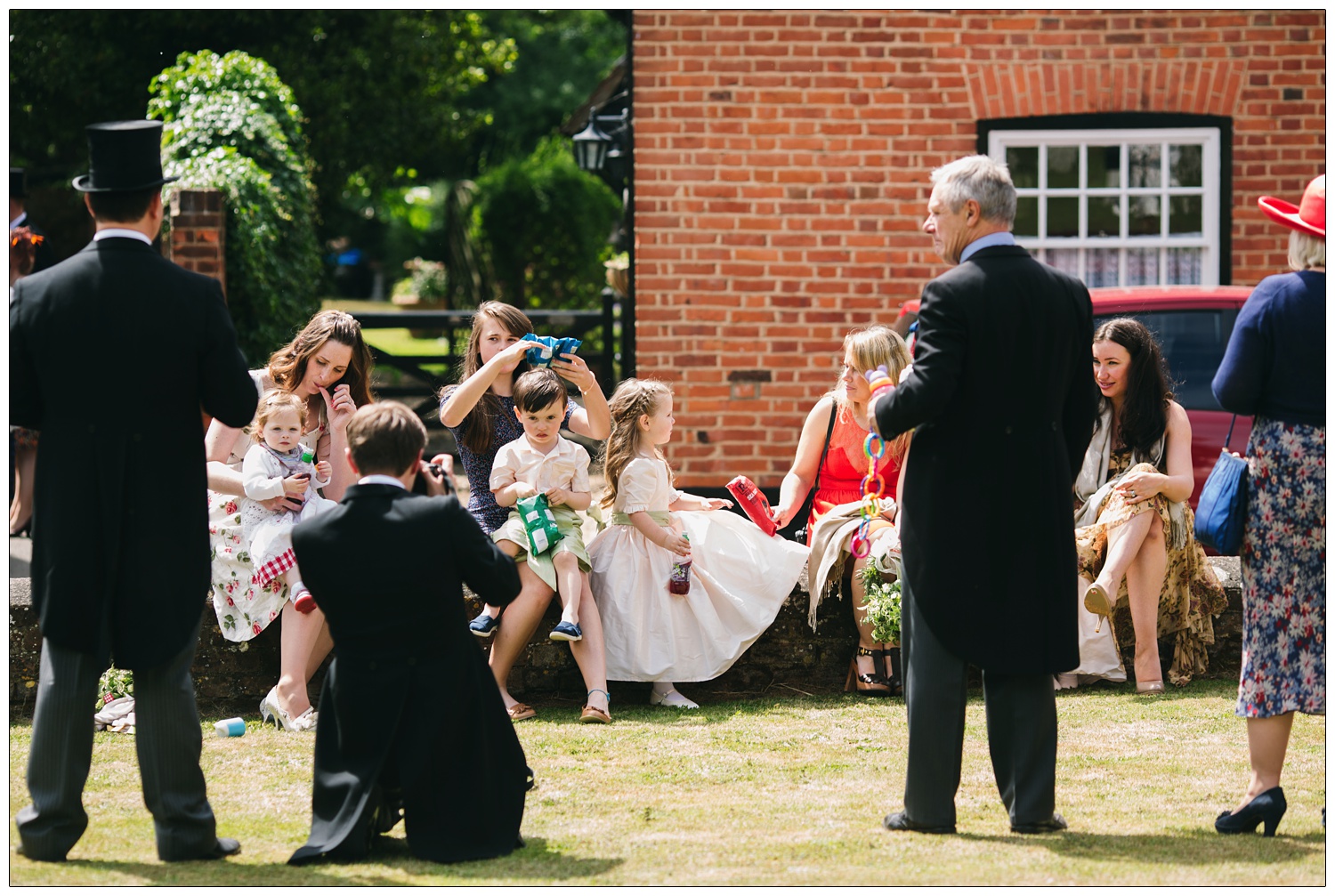 Outside the church in Bradwell-on-Sea women and children sit on a wall eating crisps. Some men from the wedding take photos of them. Wedding reportage photography.