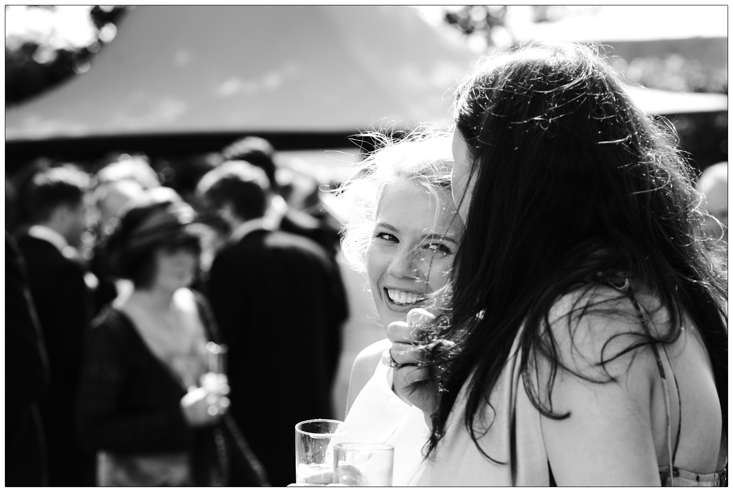 A candid wedding photograph of the bride smiling at a friend outside. They are both holding drinks.