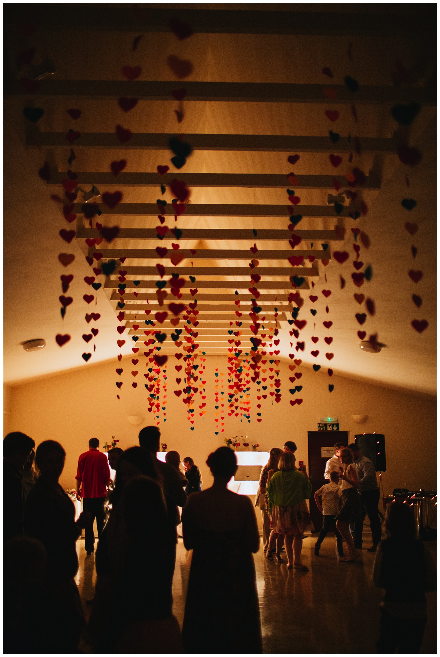 Strings of hearts hang from a ceiling. Silhouettes of wedding guests.