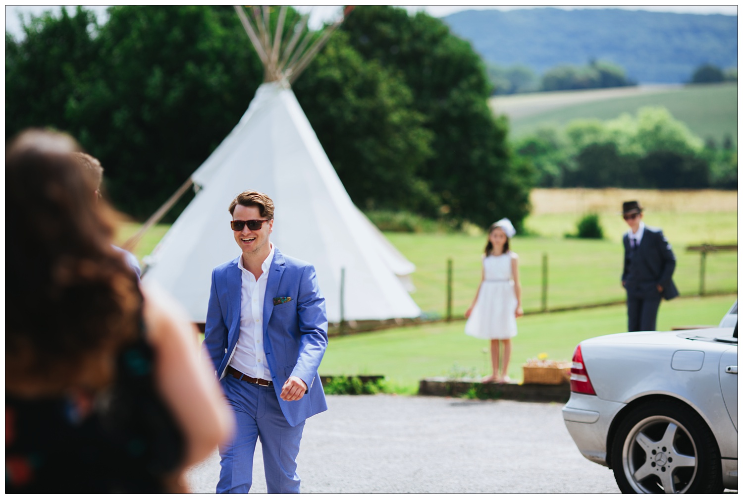 Man in blue suit and sunglasses walks in a car park. A tent and fields and trees behind him. And two children.