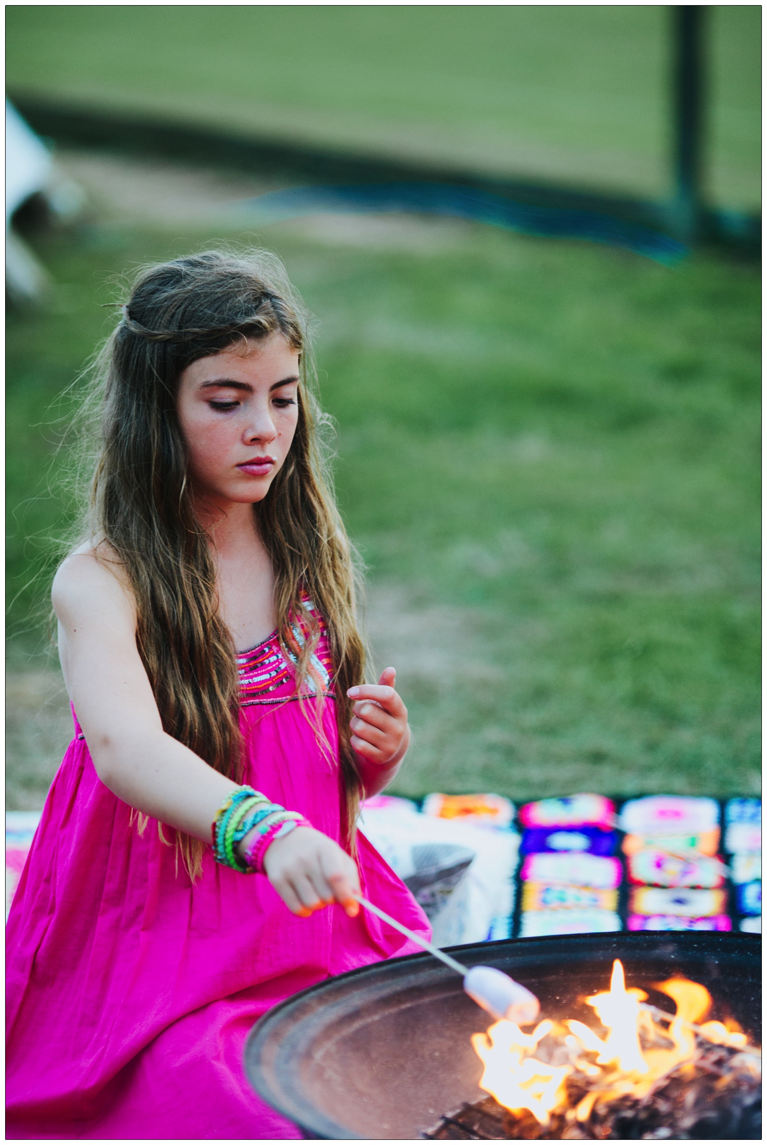 Girl in pink dress is toasting marshmallows on a fire.