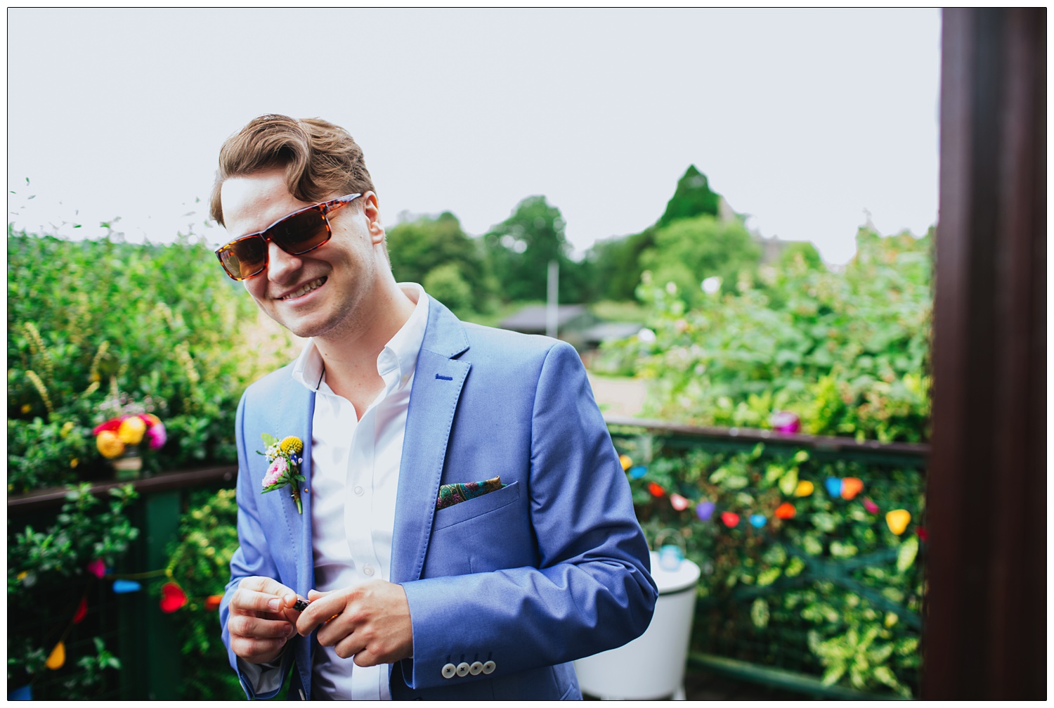 The groom in his pale blue suit and sunglasses just before his wedding. There is heart bunting on the fence.