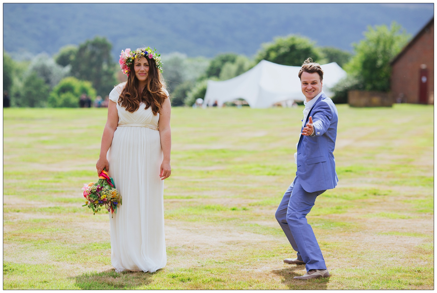 Bride is standing in her Saja wedding dress, holding colourful flowers, with a flower crown on her head. The groom is posing with his arms gesturing. He is wearing a pale blue Zara suit.