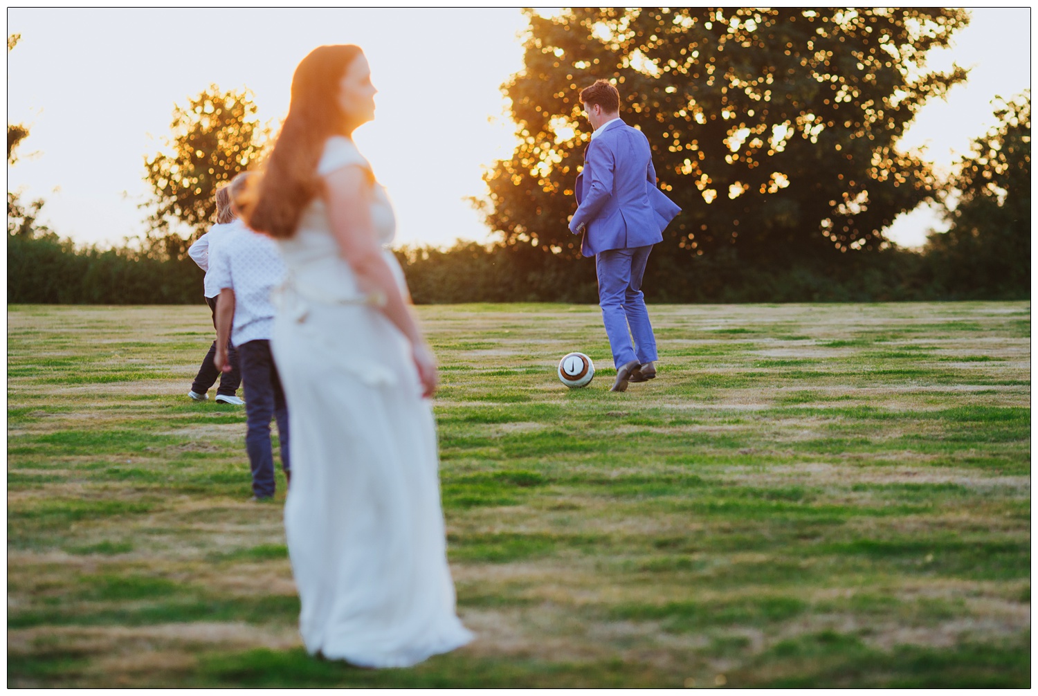 Groom in a pale blue Zara suit playing football in a field. The sun is setting and the bride is in the foreground.