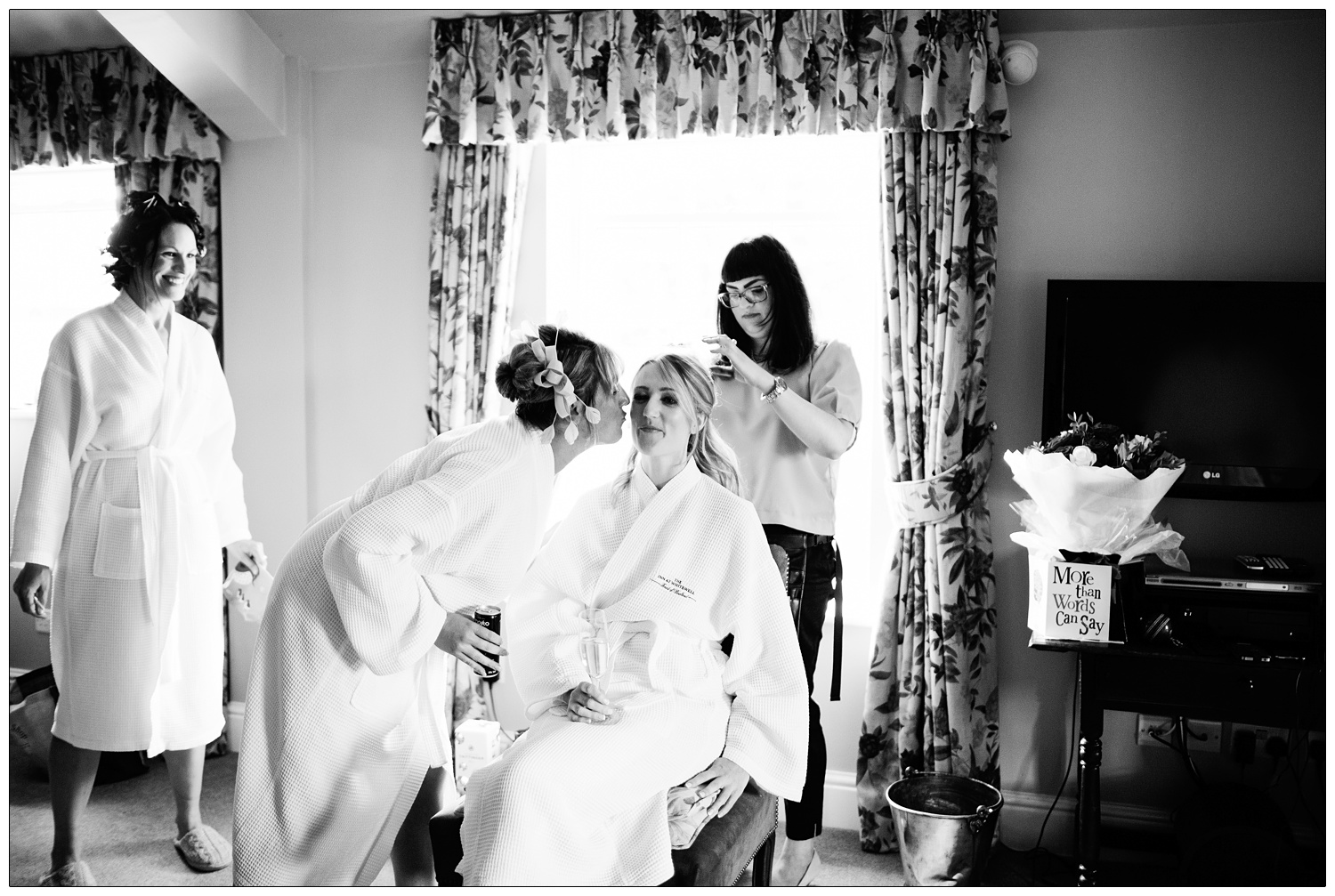 A woman goes to kiss a bride sitting on a chair. She is having her hair done.