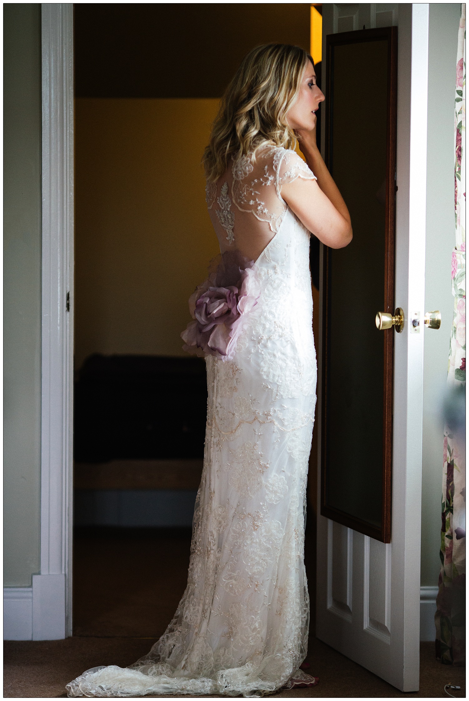 A woman in her wedding dress looks in a long mirror on the back of a door.