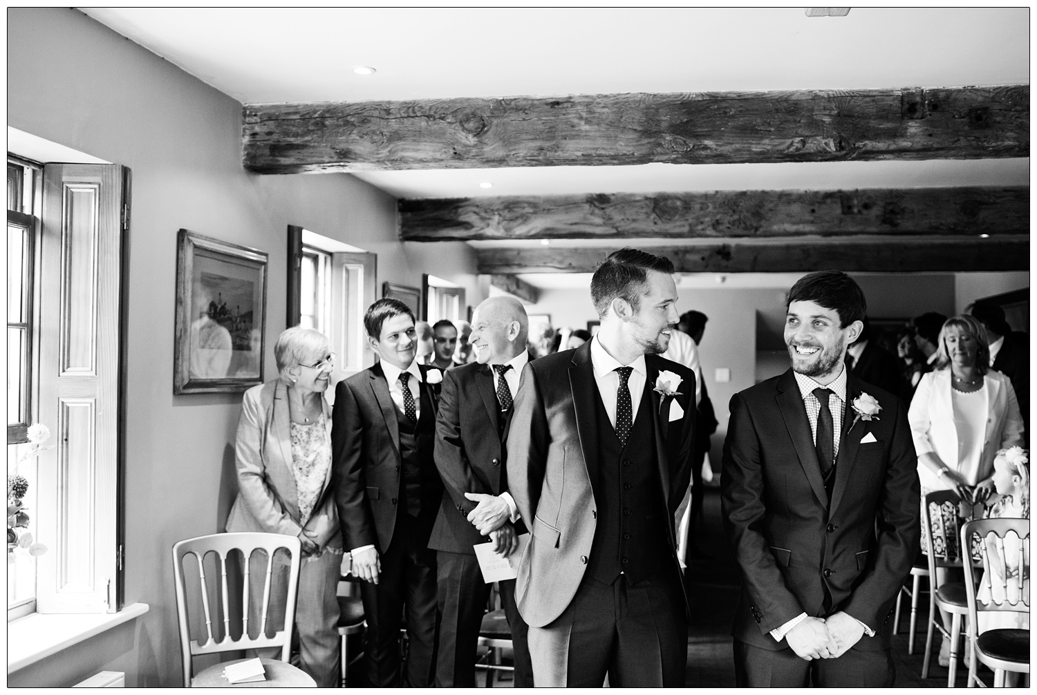 A man waits with his best man, the guests are standing up behind them in a dining room at the Inn at Whitewell.