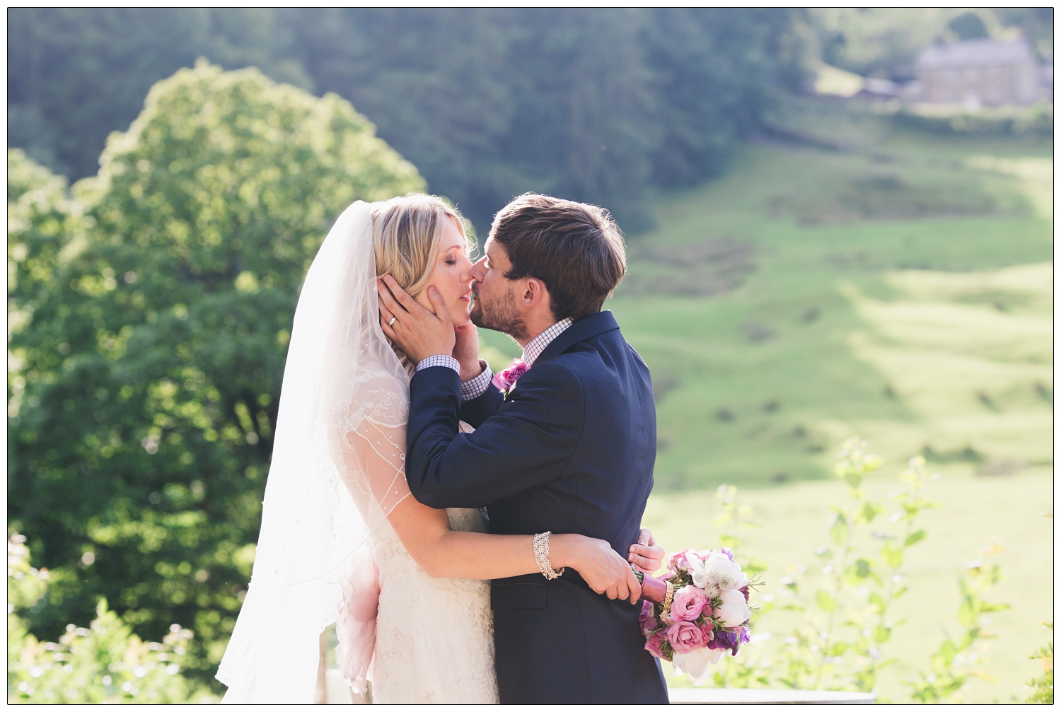 The groom holds his new wife's face before kissing her. She is wearing a veil. They are outside the Inn at Whitewell with the countryside behind them.