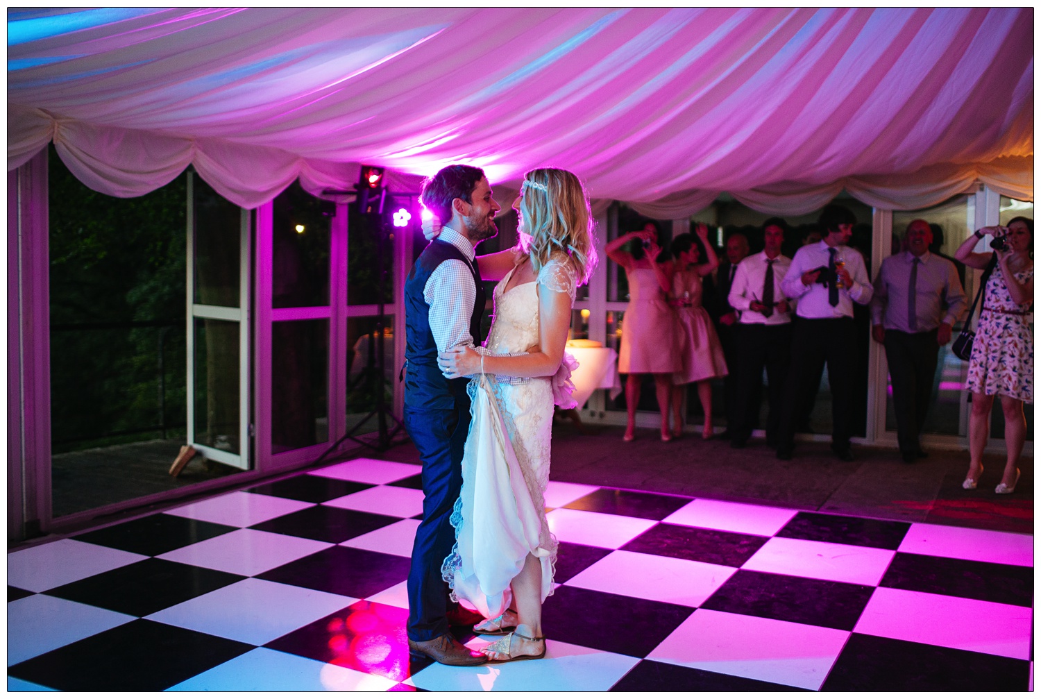 Bride and groom on the black and white chequered floor of a marquee. They are at the Inn at Whitewell having their first dance. They are lit up with magenta lights.