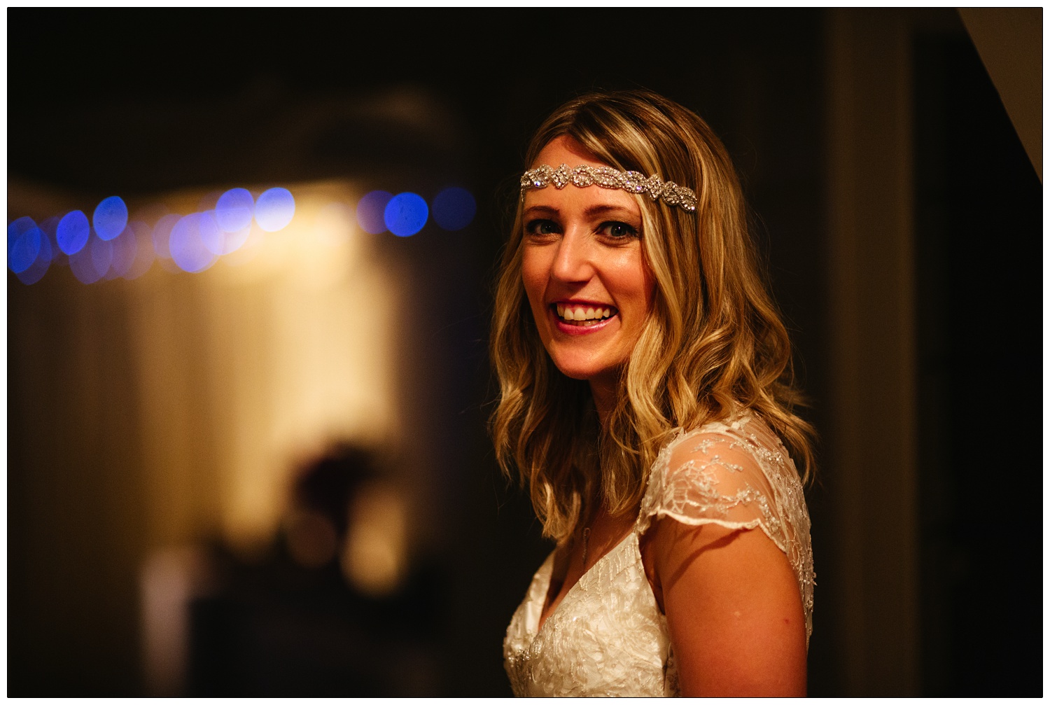The bride is a lace dress, and a headband. It's night and lighting is orange, blue fairy lights in the background. She is smiling at the camera.