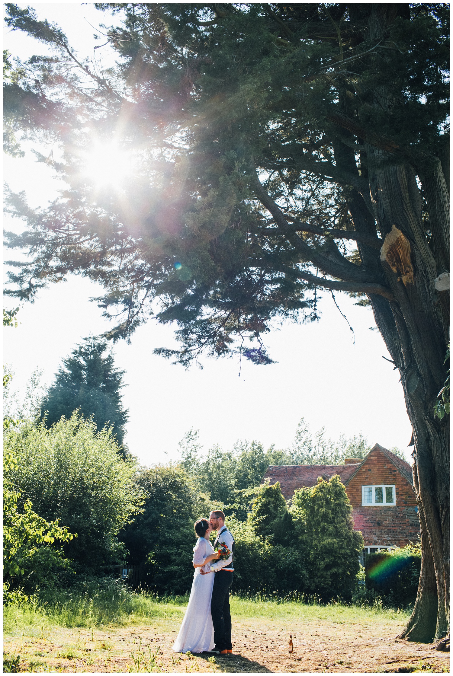 A newly married couple kiss under a tall tree at The Kench Hill Centre. The sun is flaring through the branches.