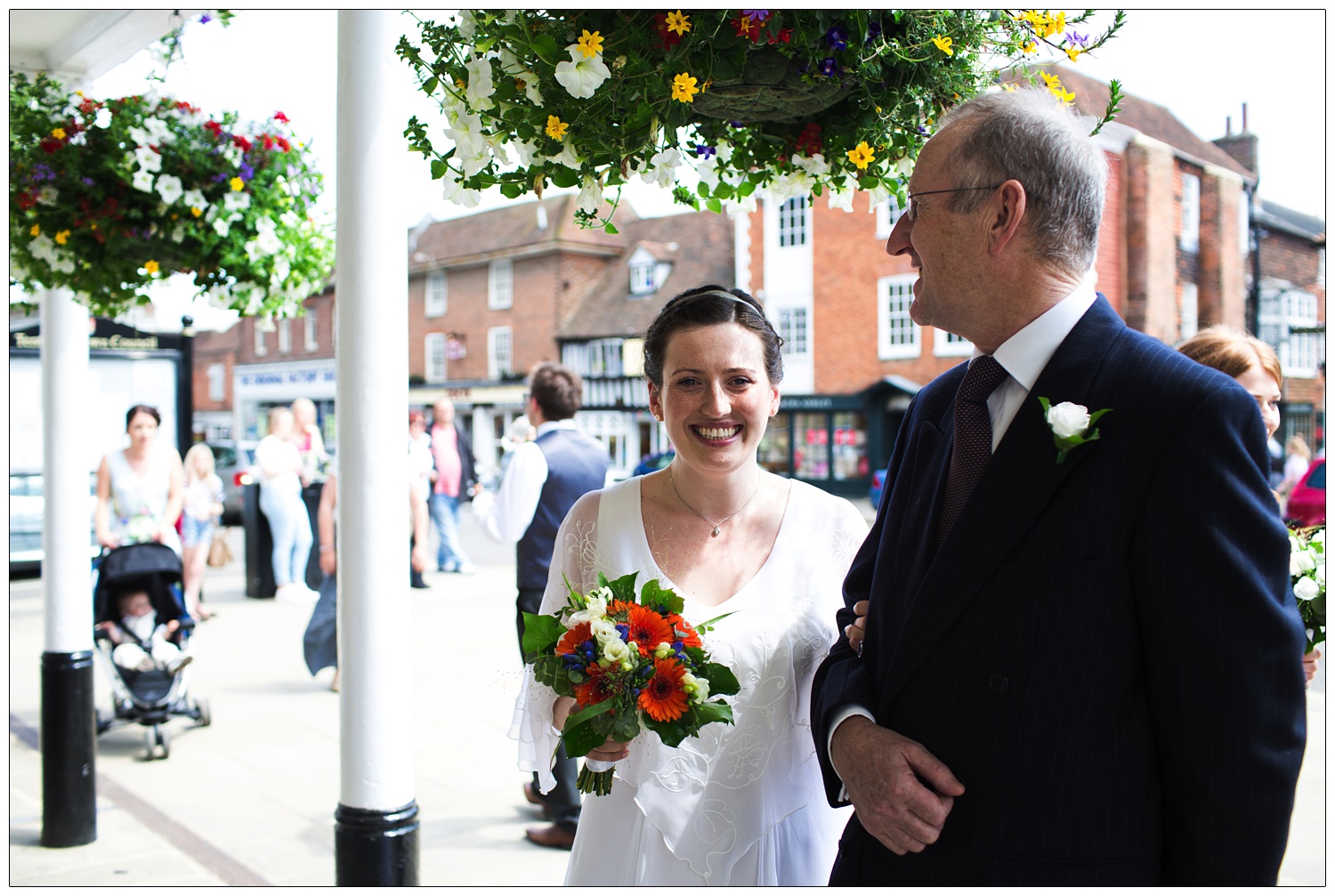 A woman in her wedding dress with her dad outside the town hall in the Tenterden. There are hanging baskets full of flowers.