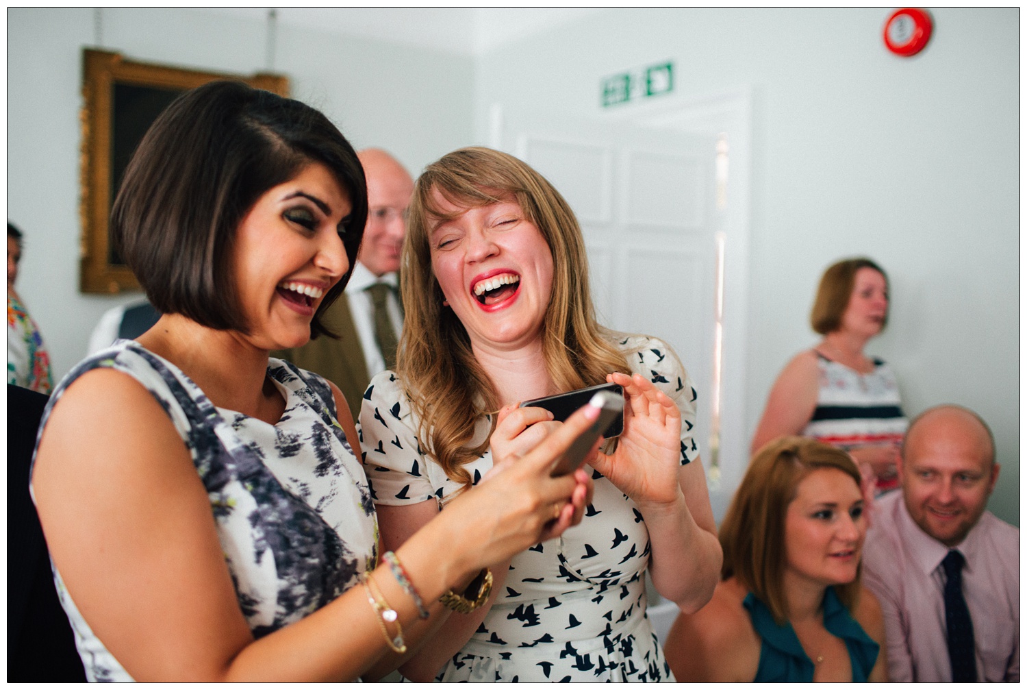 Two women laughing at their phones in the wedding ceremony room at Tenterden town hall.