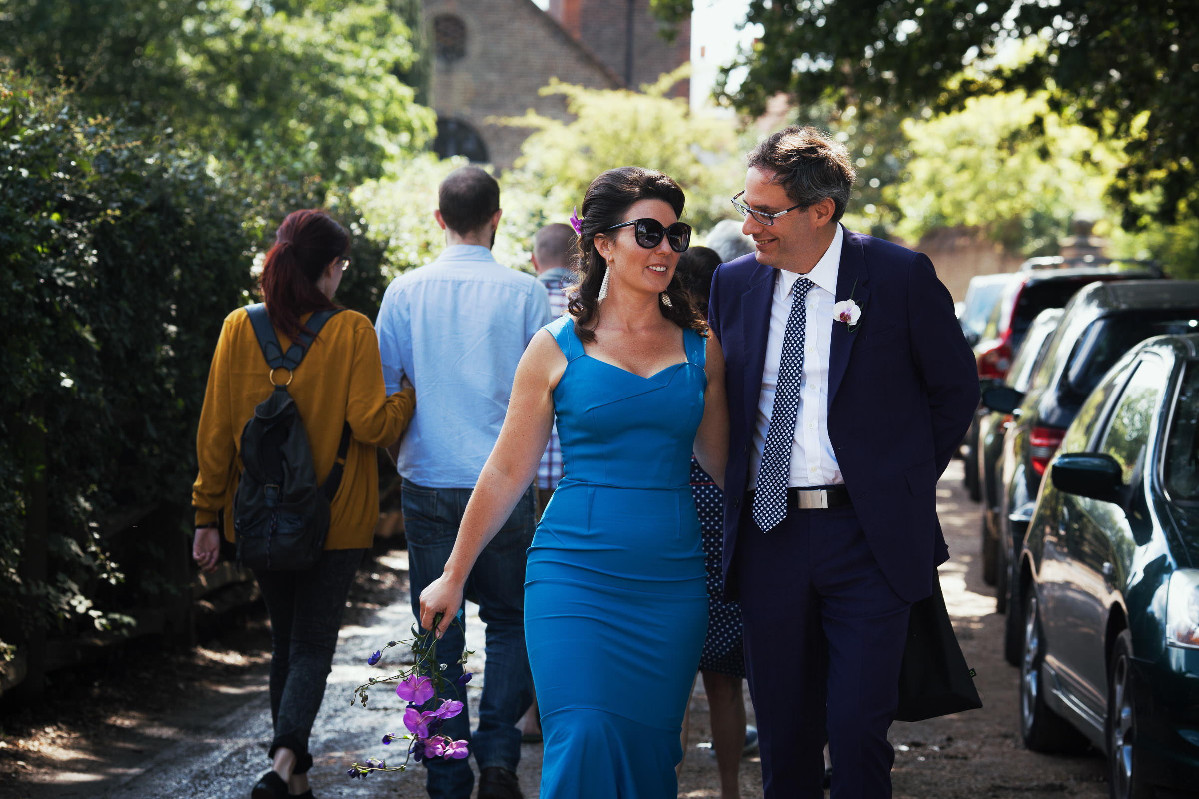 Bride and groom walking off Church Lane to Petersham Nurseries. Cars parked on the right. The woman is wearing Roland Mouret Mermaid Dress in Blue and sunglasses. The groom is wearing a purple suit. It's a sunny day.