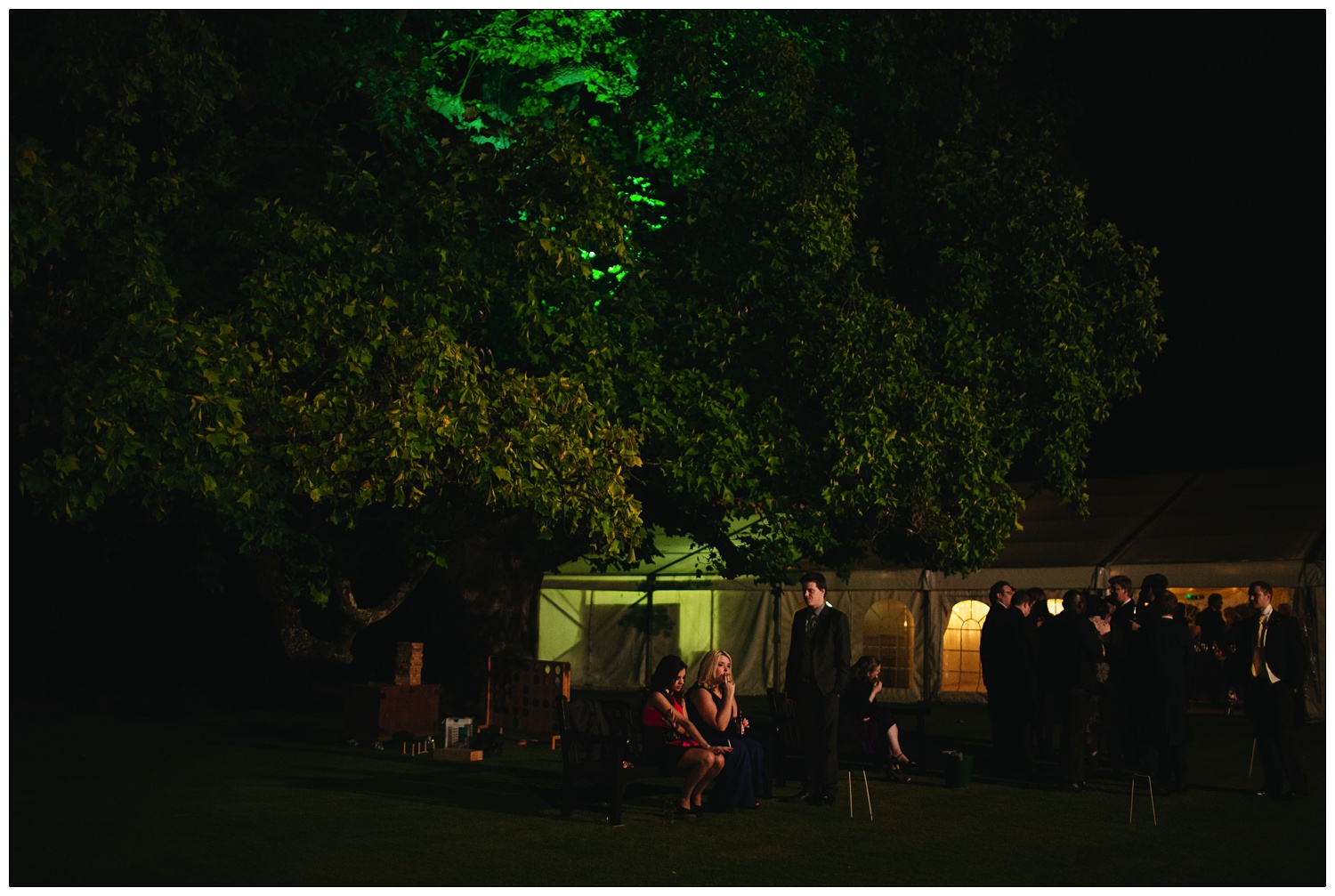 People sat on a bench at night under a tree. There is a marquee in the background. They are at a wedding reception at Hedingham Castle in Essex. It's reportage style wedding photograph.