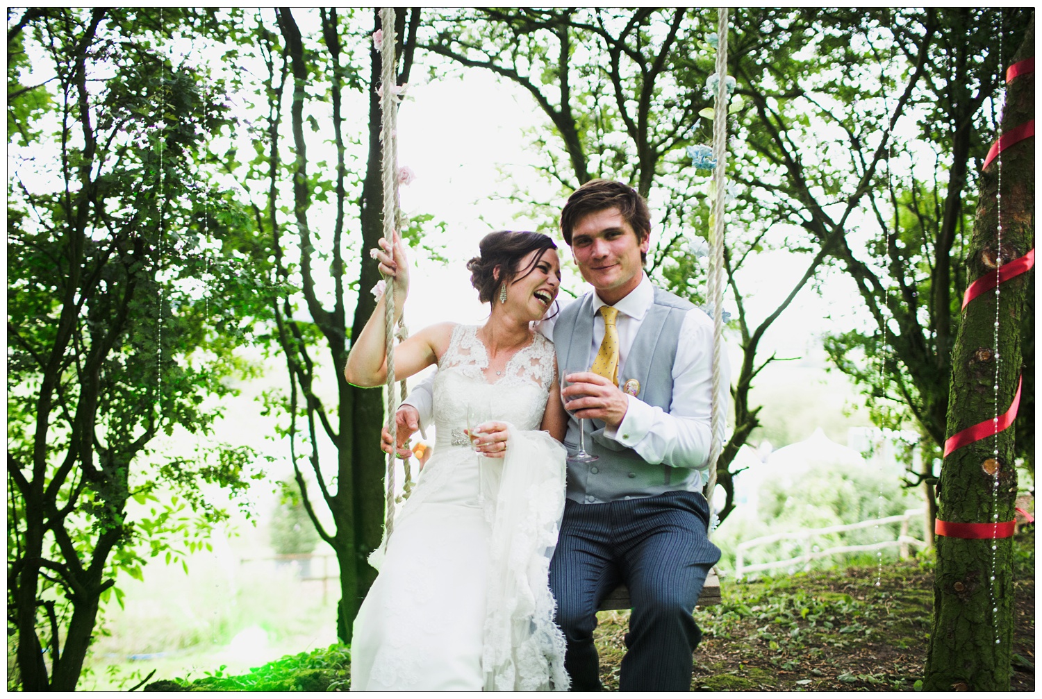 Bride is laughing and looking at the groom. They are sat on a swing in the trees holding wine glasses..