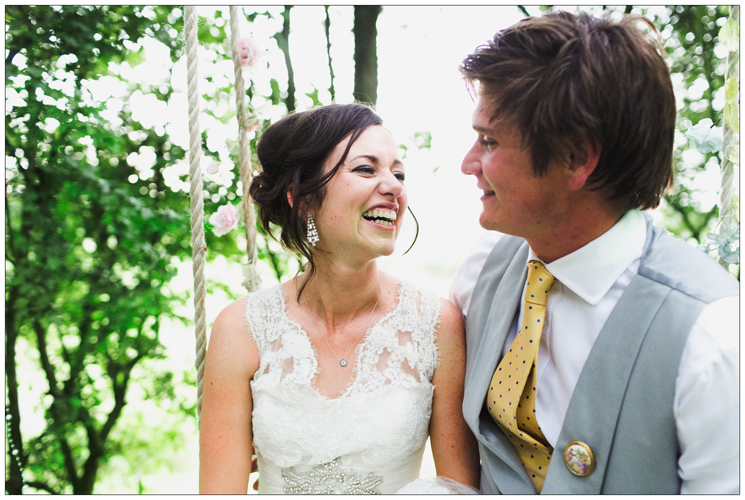 Bride is laughing and looking at the groom. They are sat on a swing in the trees.