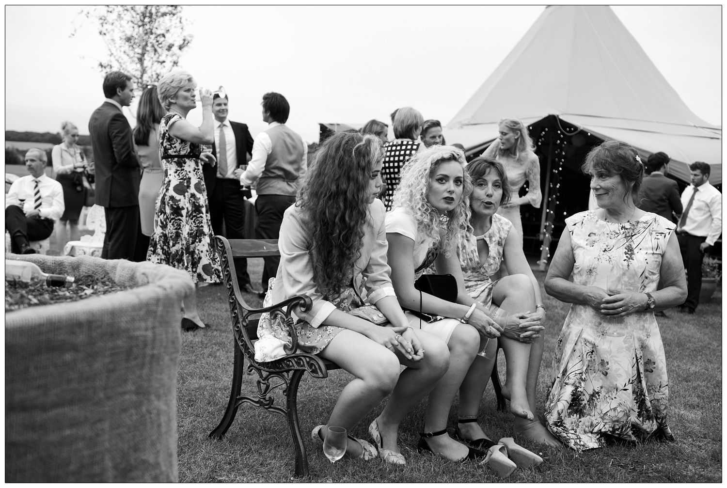 Three women on a bench, one woman kneeling on the grass at a wedding. ONe woman has taken her shoes off. There is a Tentipi behind them.