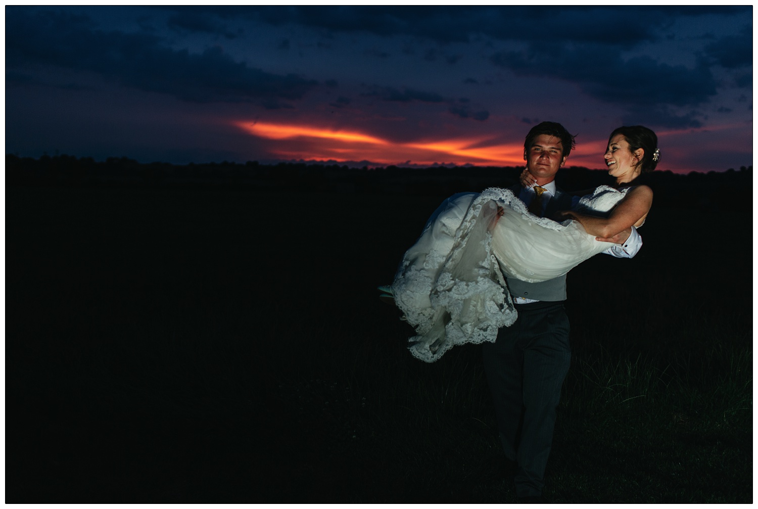 The sun has recently set. The sky is orange and purple. A man is carrying a woman in a wedding dress.