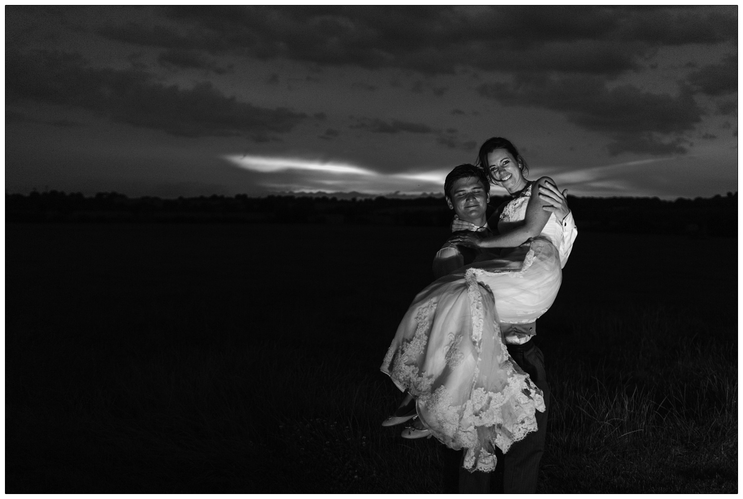 Black and white photograph taken at twilight. The groom is carrying his new wife.