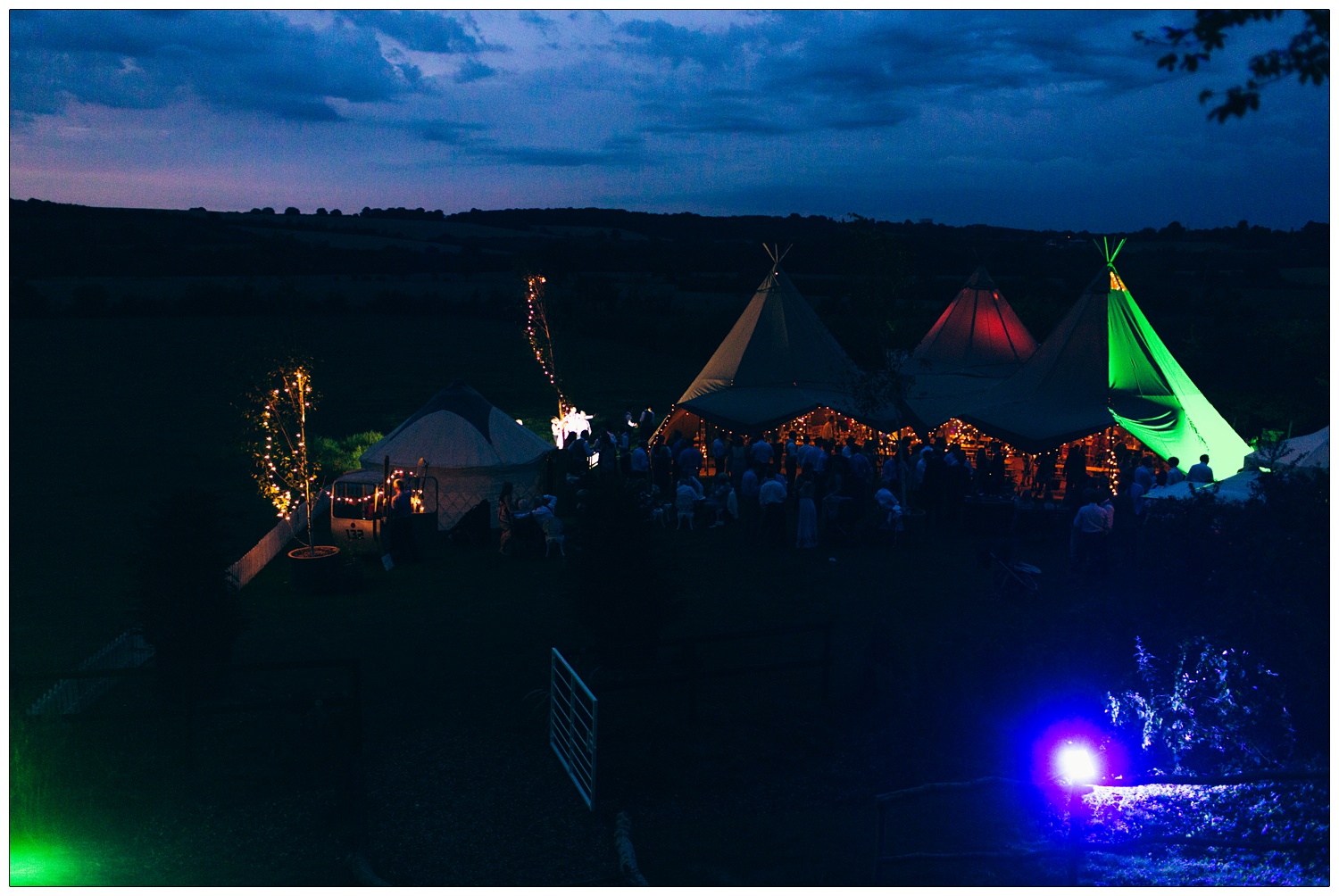 A twilight view from above over a wedding at home on a farm. there is a ski cable car, a yurt and a Tentipi. There are red, green and blue lights. There are strings of lights. Fields in the background.