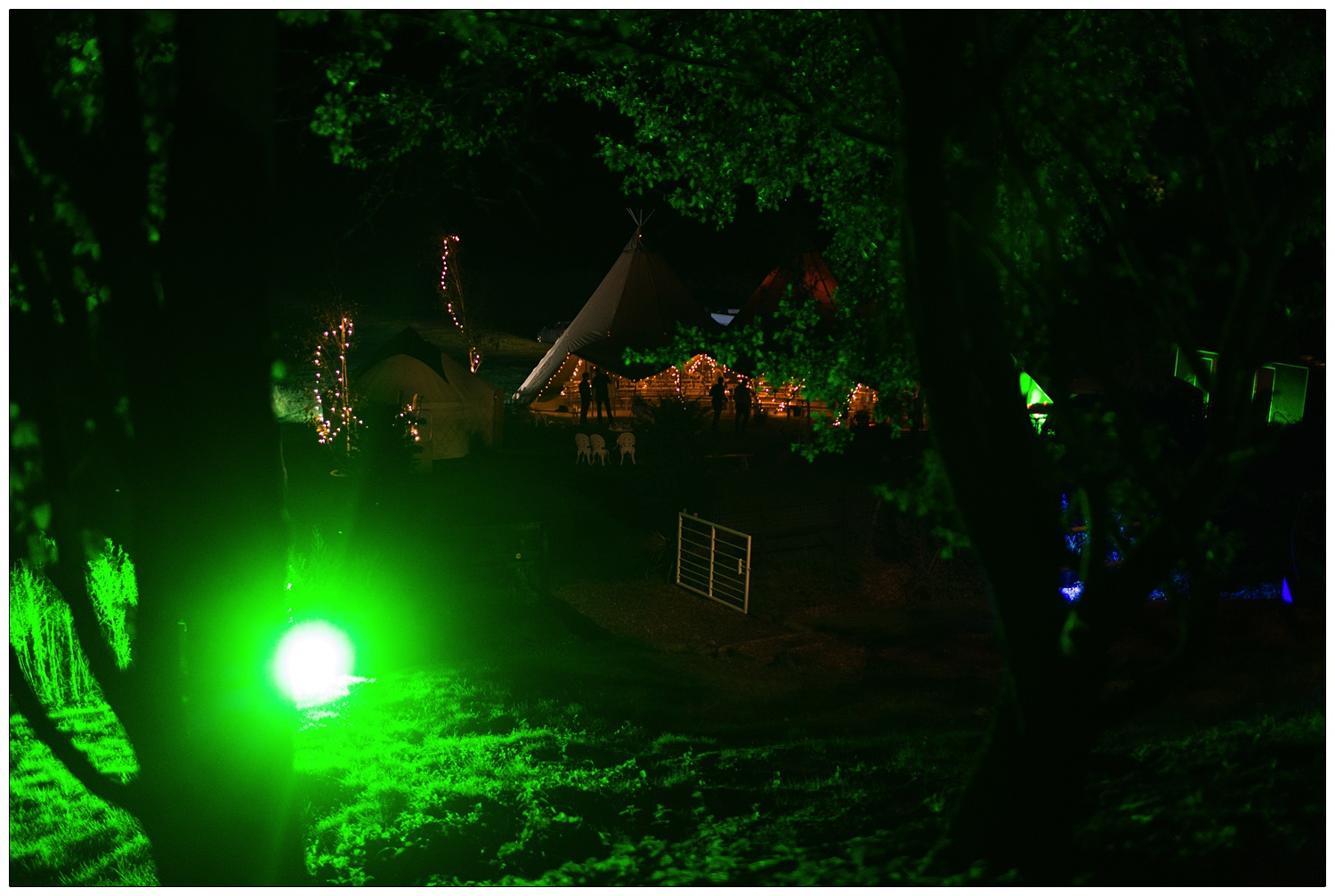 Up a hill looking down the view of a festival themed wedding. A tipi and yurt, there is green lighting by a tree.