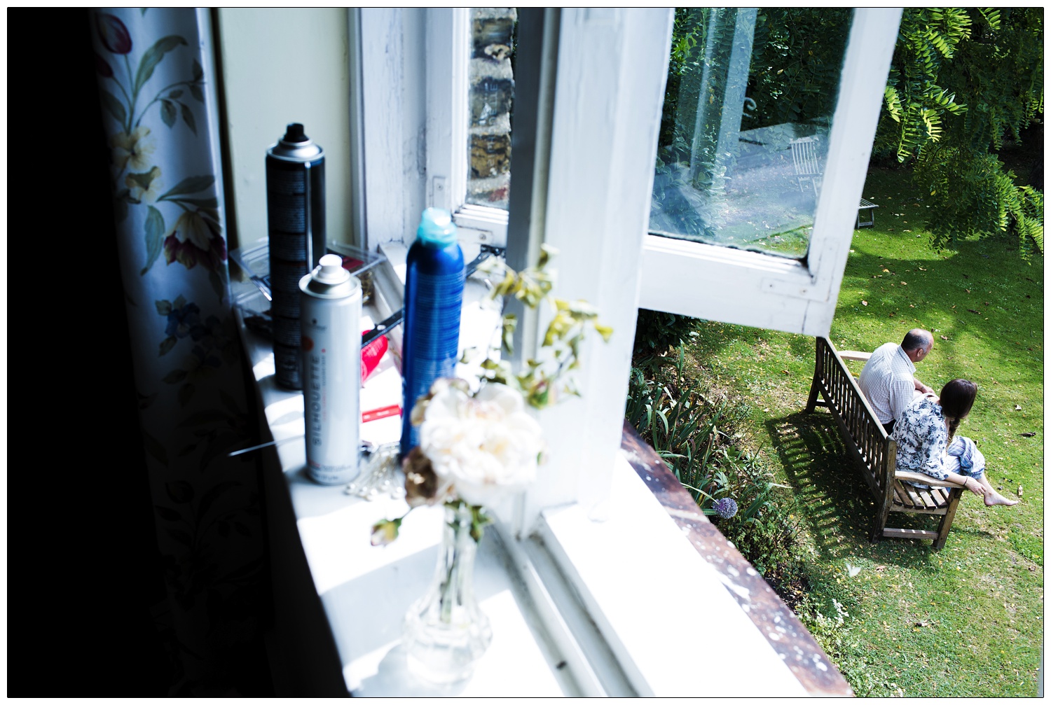 Looking out of a bedroom window to see a man and his daughter sat on a bench in a garden.