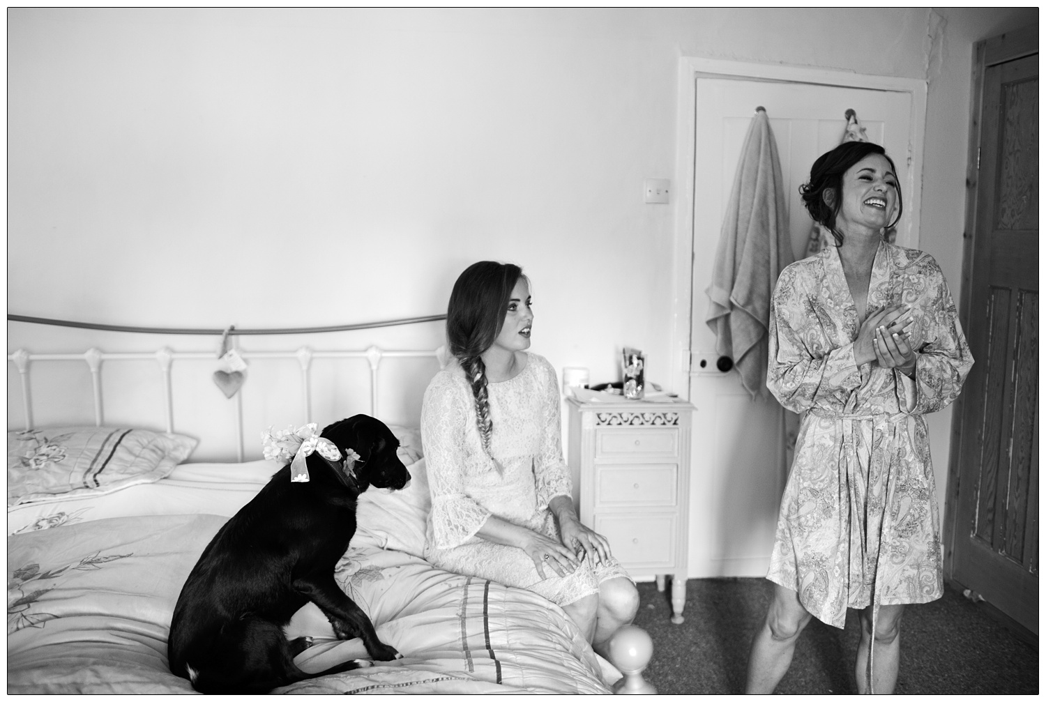 Bride is in a dressing gown with her sister, a dog sits on the bed.
