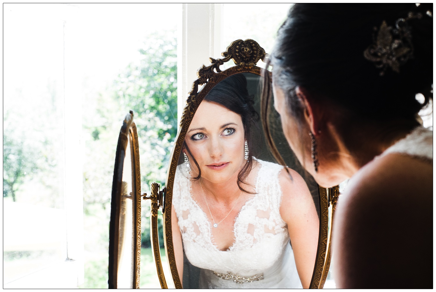 A bride looks in a mirror on a dressing table in front of a window.
