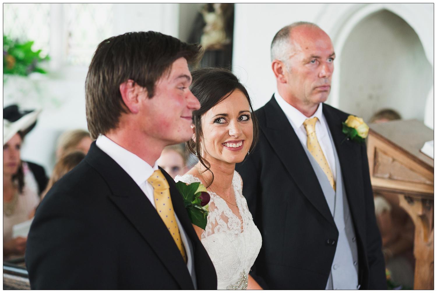 The bride is stood between the groom and her dad. She is looking at her husband-to-be in a church in Essex.