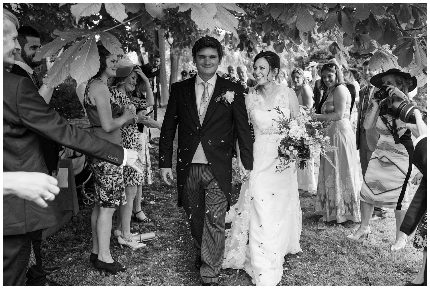 In the grounds of The Church of St Mary & St Margaret in Stow Maries a newly married couple walk down the path. Under the trees. People are throwing confetti.