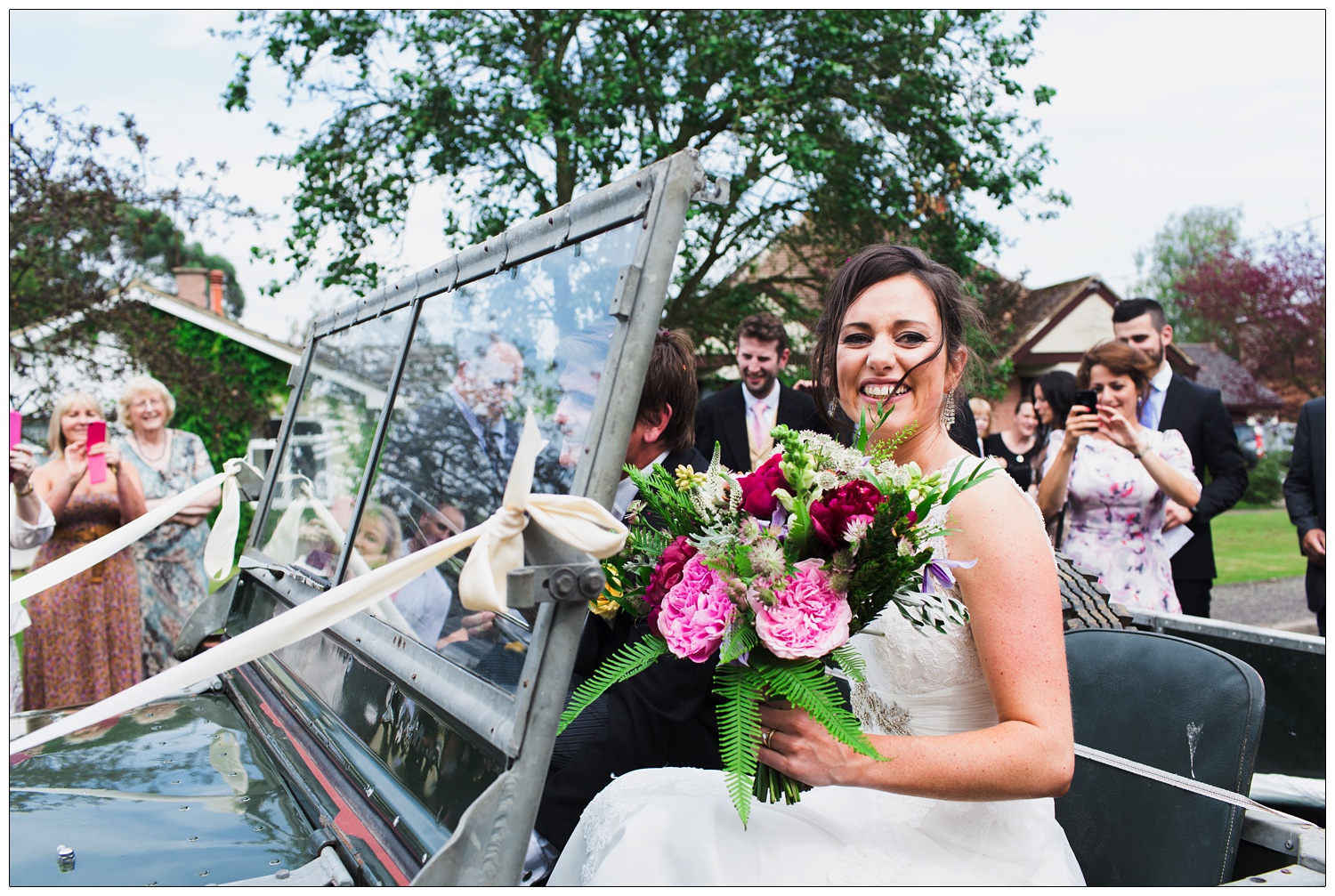 Bride is smiling and holding flowers. She is sat in a Land Rover and her husband is driving.