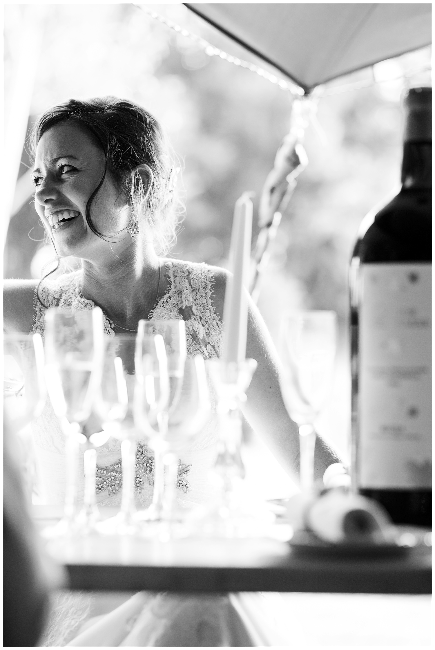 A reportage style wedding photograph of a bride laughing sat at a table. The table is full of champagne flutes and a big bottle of wine.