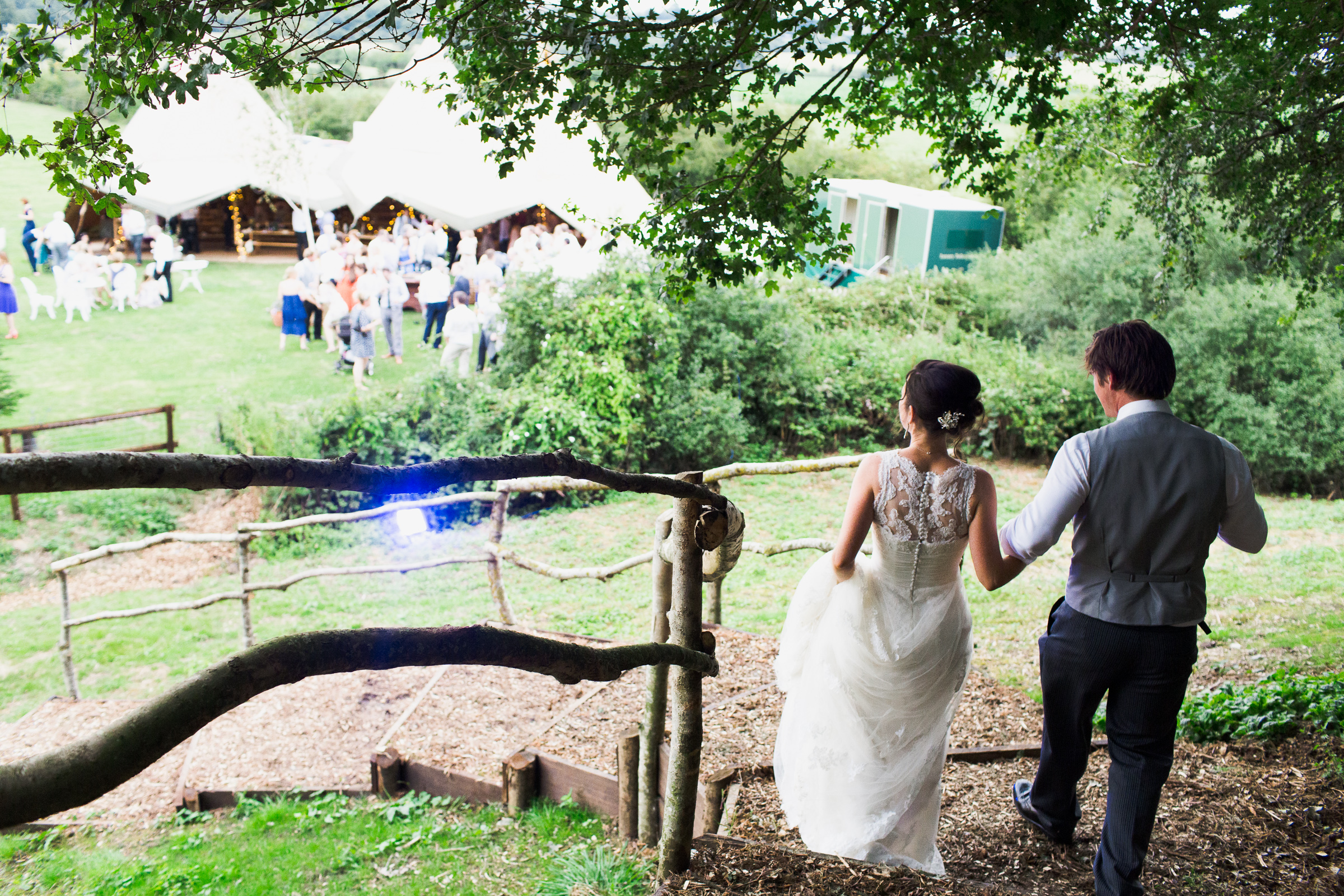 View from behind of bride and groom walking down the wooden steps to their wedding reception in a field. There is a tipi in the field and wedding guests. There are trees above them.