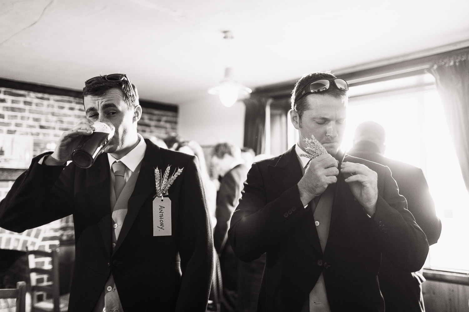 Groomsmen in the The Green Man Inn in Bradwell-on-Sea. One man is drinking a pint, the other is fixing his buttonhole, which is made of ears of wheat. They have sunglasses on their heads.
