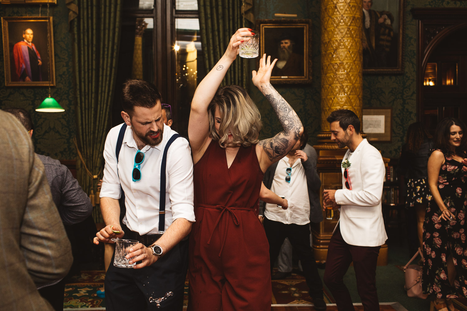 Two people dancing at a wedding reception at The National Liberal Club. Some drink has spilt out of the man's glass and is in the air. The woman is wearing a dark red jumpsuit.