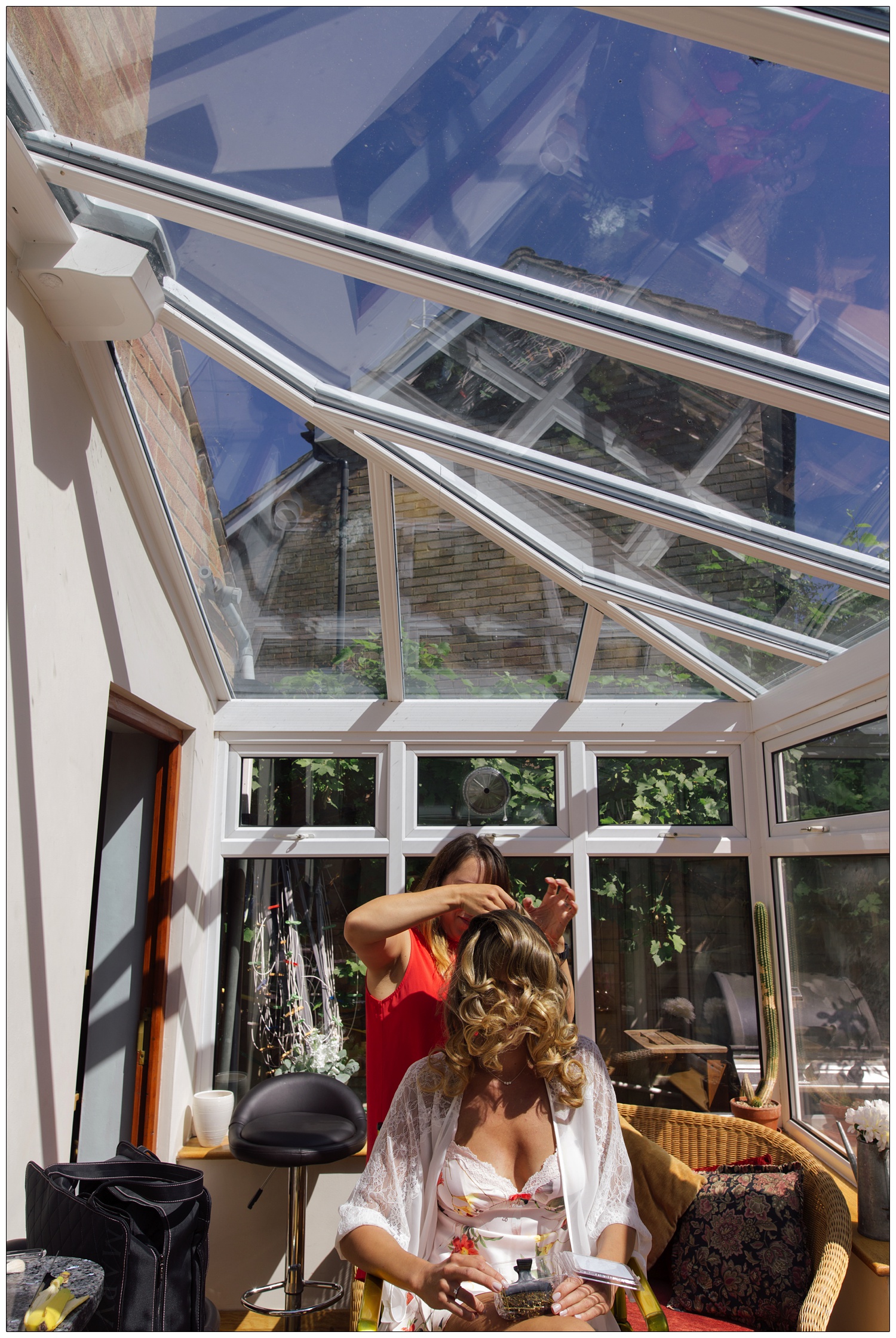 In a conservatory at home a woman is having her hair done. The sky is blue.