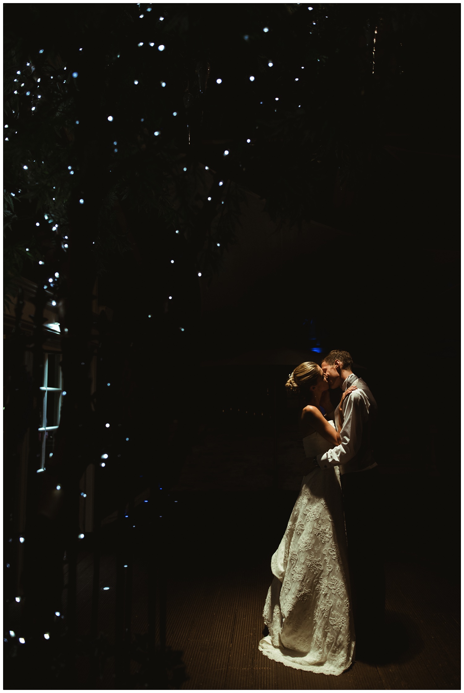 A newly married husband and wife kiss in the dark. There are lights in a tree at Friern Manor