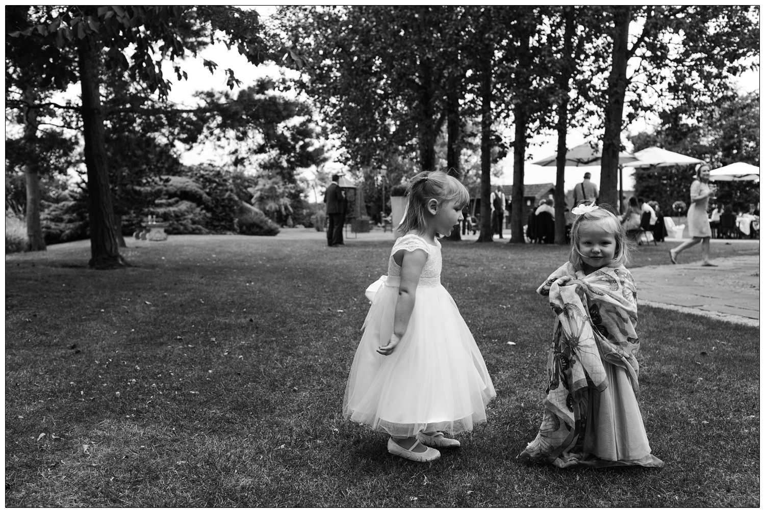 Two small girls in the grounds of Friern Manor. One girl is in a white dress, another is wrapped up in a scarf. There are trees in the background, and wedding guests drinking.
