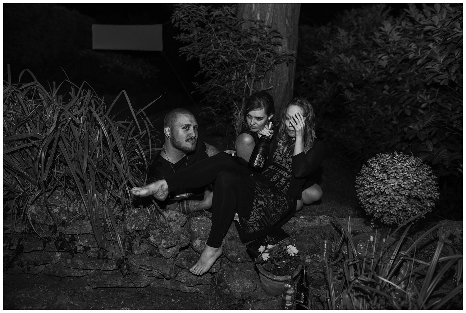 A man and two women are drinking from a champagne bottle on the floor in a garden. A reportage style wedding photograph.