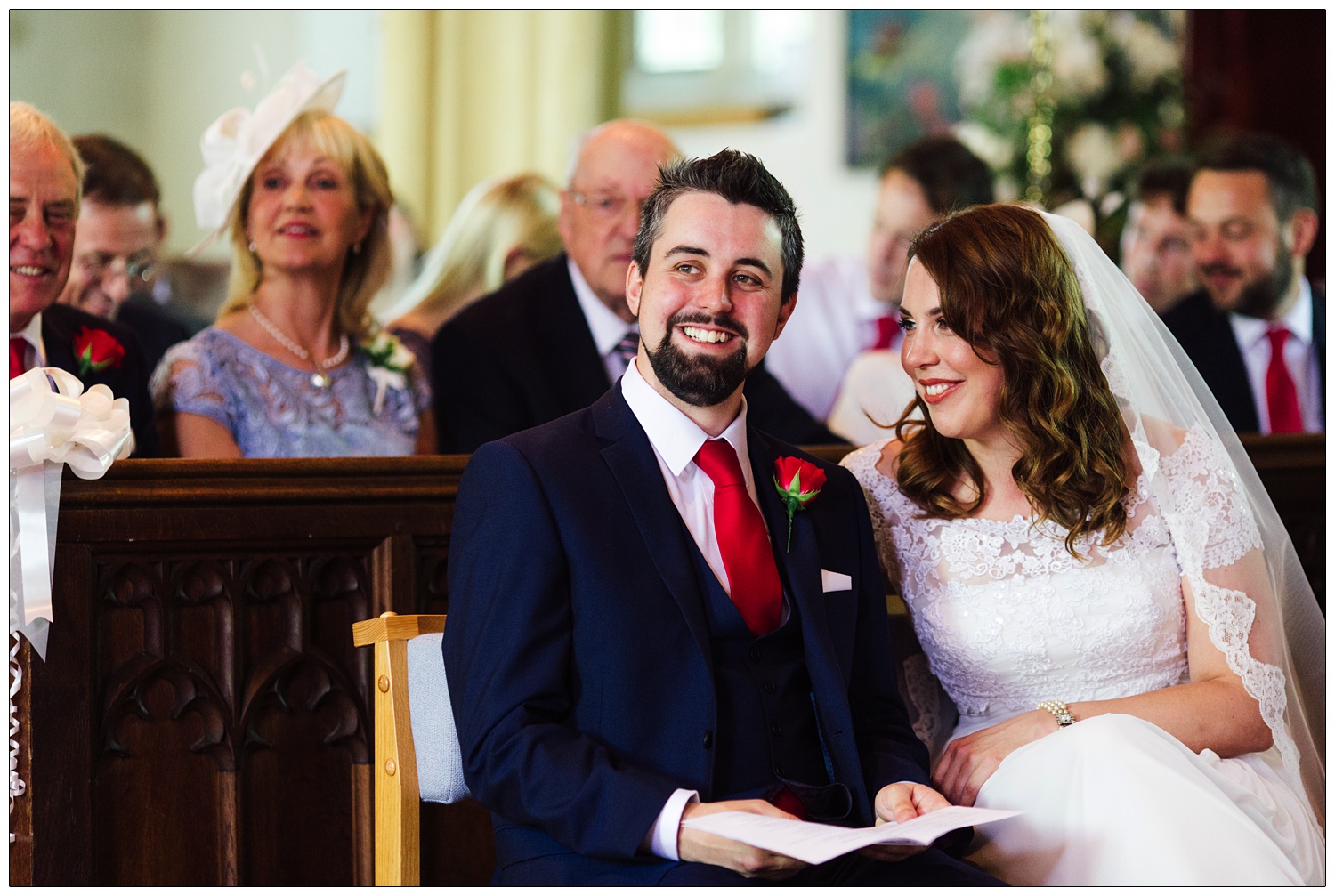 Bride and groom sat in chairs at the wedding ceremony at St Andrew's Church, Chew Magna. The bride is looking at the groom. He is in a dark blue suit with a red tie. She is wearing a lace boatneck dress.